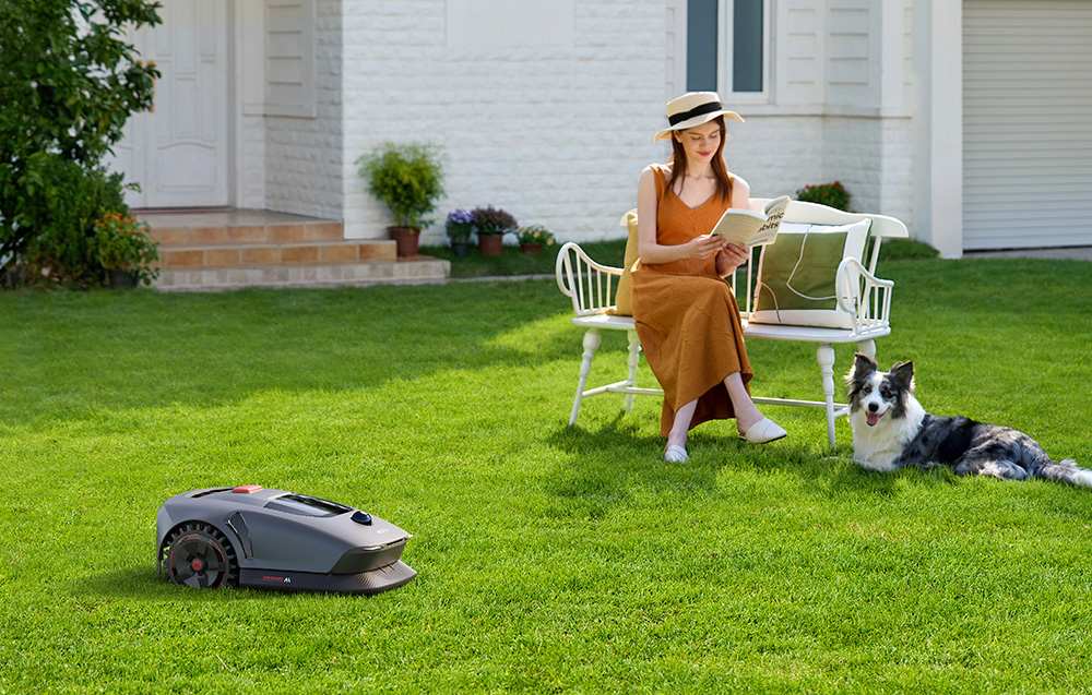 Na bílé lavičce sedí tmavovlasá žena a čte si knihu. Vedle ní leží pes. Nalevo seká trávník robot.